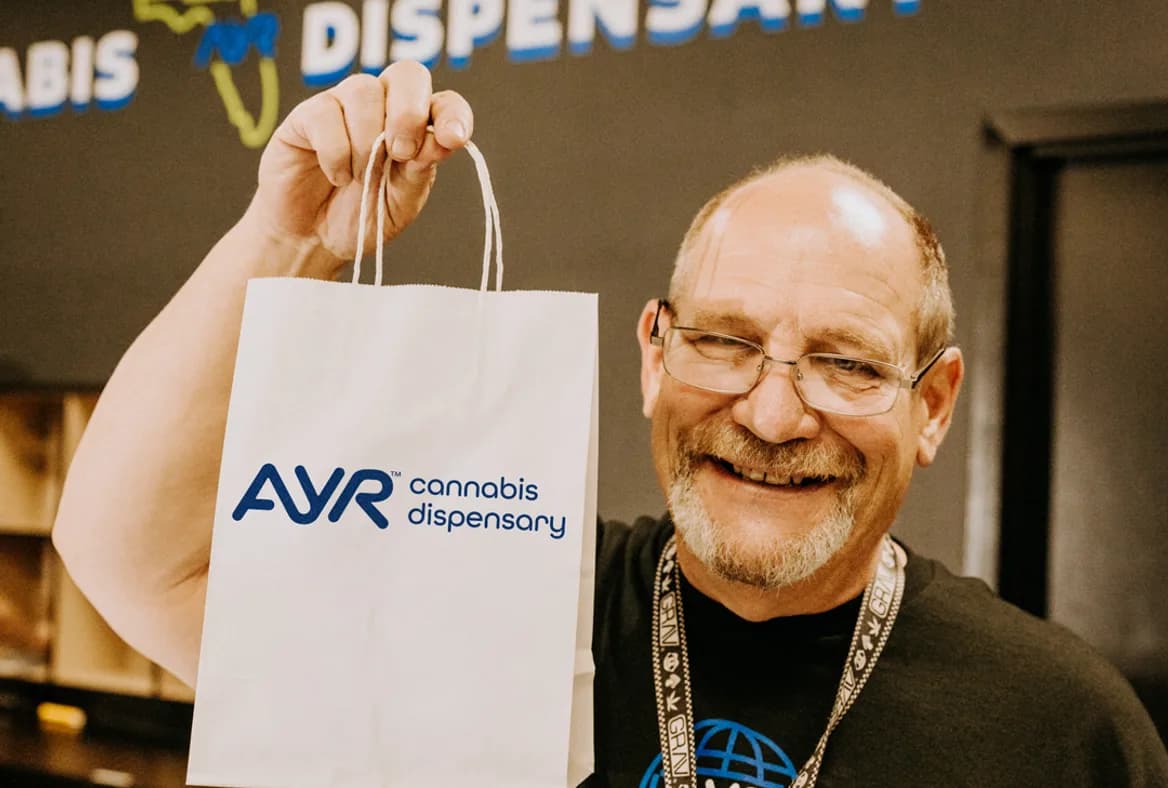 Smiling Ayr Dispensaries customer holding an Ayr dispensary bag at the counter